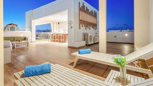 A rooftop lounge area with wooden flooring, white lounge chairs, and a bar area in the background. - NEW 2BR Condo  Pool, Gym, & Steps from Palm Beach (Palm Beach)