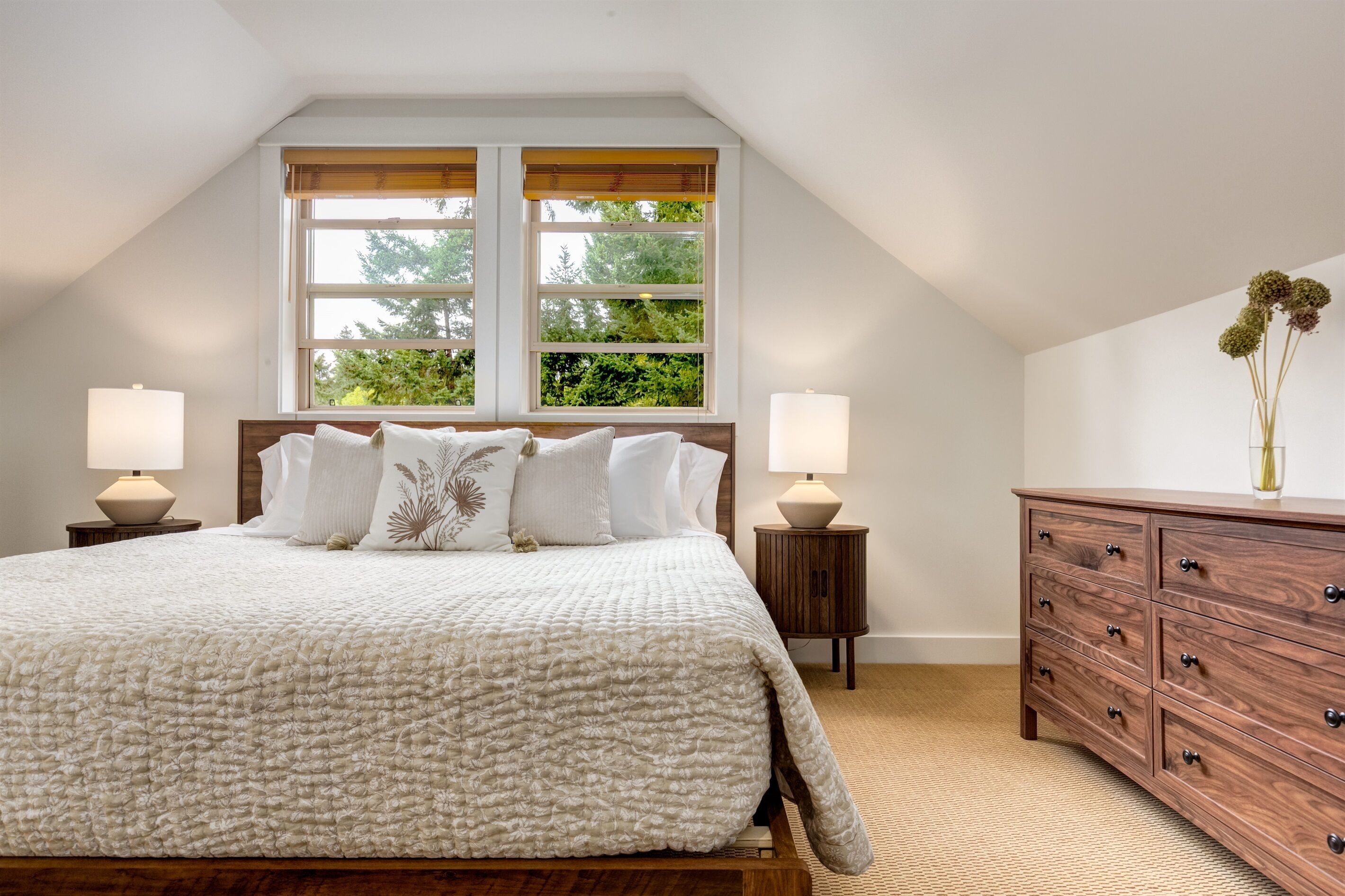 A bedroom with a large bed, two bedside lamps, a wooden dresser, and a window with a view of greenery.