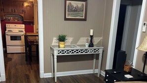 Een woonkamer met een bruine bank, een gekleurd vloerkleed, een witte tafel met een plant en een kaars, en een keuken op de achtergrond.