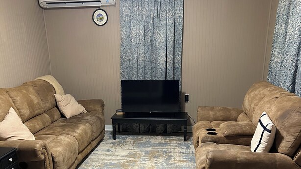 Een woonkamer met twee bruine banken, een flatscreen televisie op een standaard, een tapijt met een patroon, houten vloeren en een airconditioning op de muur.