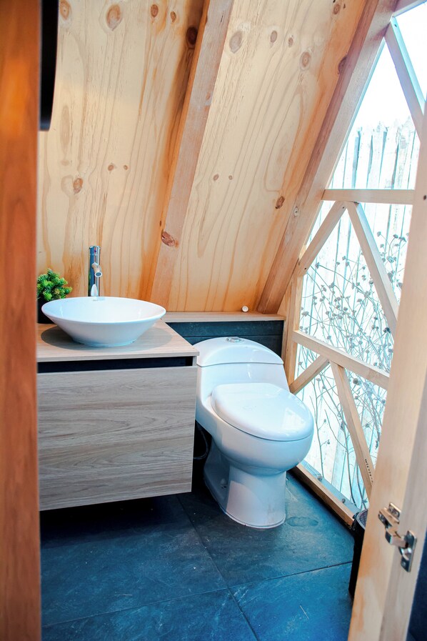 Un baño de madera pequeño y acogedor, con un lavamanos y un inodoro blancos, y una ventana con marco de madera.
