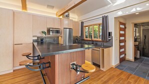 A modern kitchen with wooden cabinetry, a central island with stools, stainless steel appliances, and a window overlooking greenery.