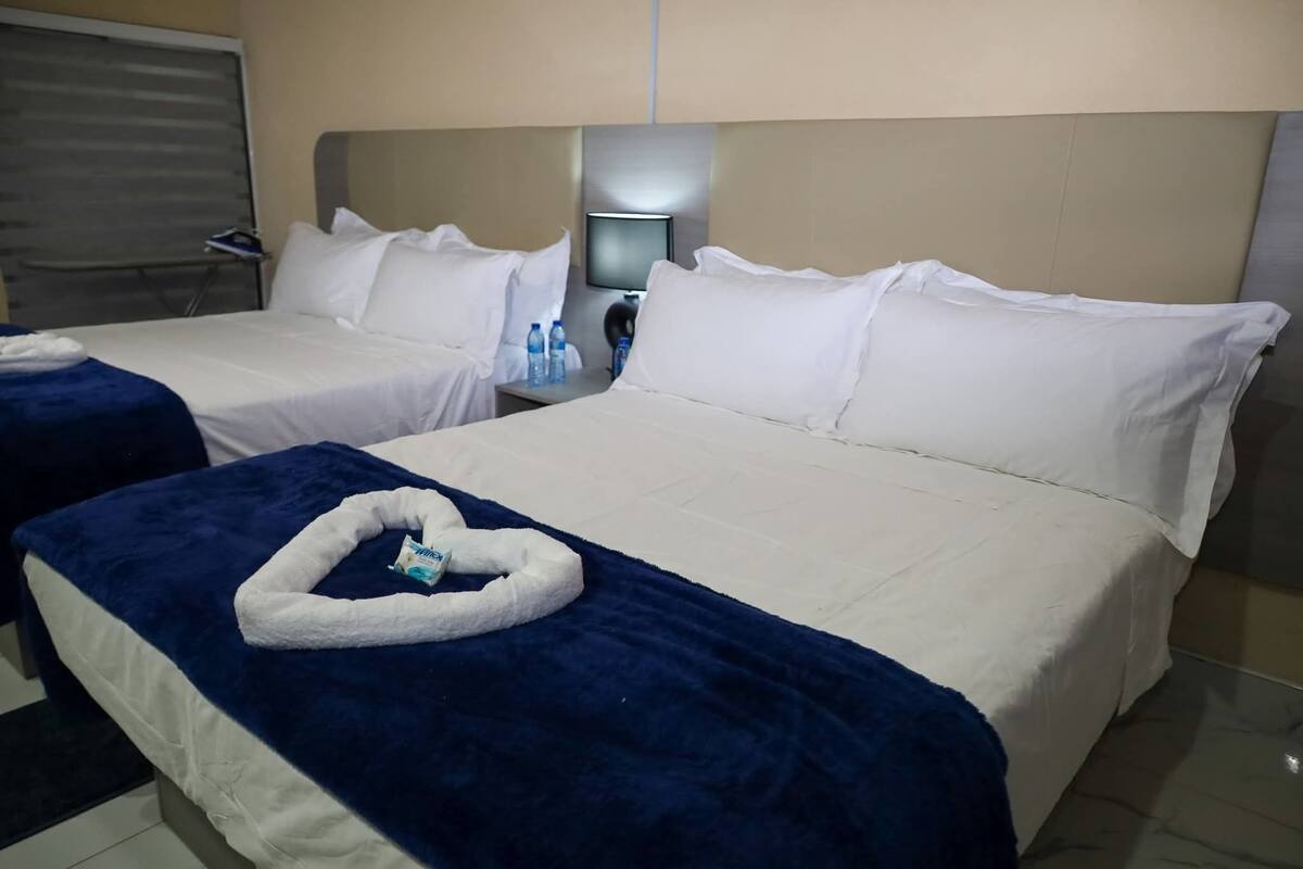 A hotel room with two beds, each with white linens and a blue blanket. There is a small table with a lamp and a bottle of water.