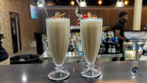 Two tall glasses of a creamy beverage with garnishes, placed on a bar counter in a restaurant with a brick wall and hanging lights in the background. - CROWN HOTEL GUYANA INC (Georgetown)