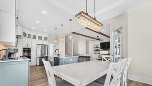 A modern kitchen with white cabinetry, a large island, and a dining area with white chairs. - Brand New 7 Bedroom, 7 Bathroom Beach House (Ocean Isle Beach)