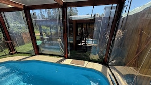 Una piscina con cerramiento de vidrio, rodeada por una terraza de madera y vista al jardín.