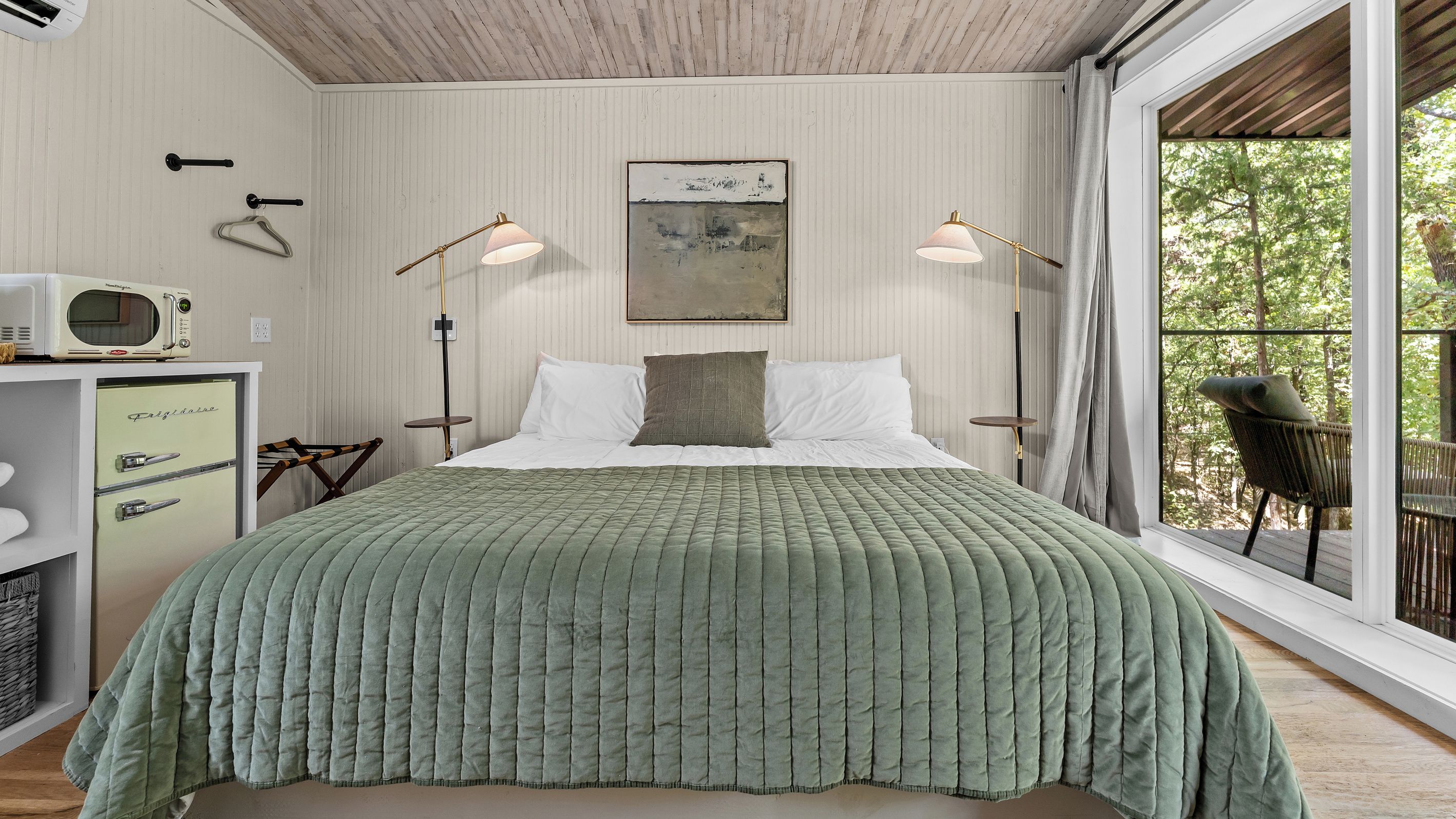 Un dormitorio con cama, lámparas de noche, un cuadro, un refrigerador antiguo y vistas a árboles a través de una ventana.