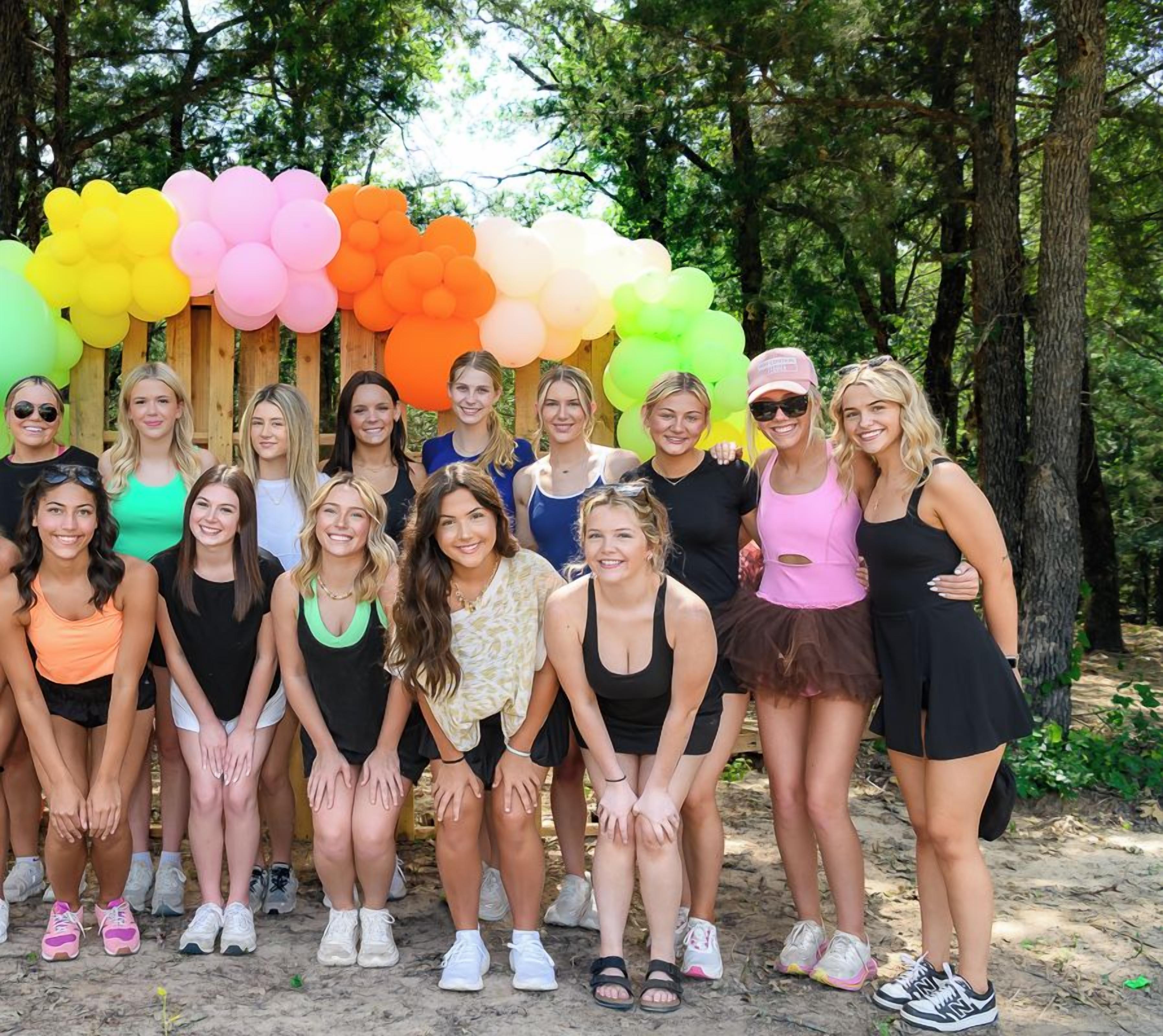 Een groep mensen poseren voor een kleurrijke ballonboog in een bosrijk gebied.