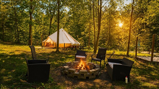 Ein Zelt im Wald, eine Feuerstelle mit Stühlen und eine Bank.