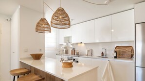 Une cuisine moderne avec des meubles blancs, un îlot central doté de tabourets, des suspensions en osier et une planche à découper en bois.