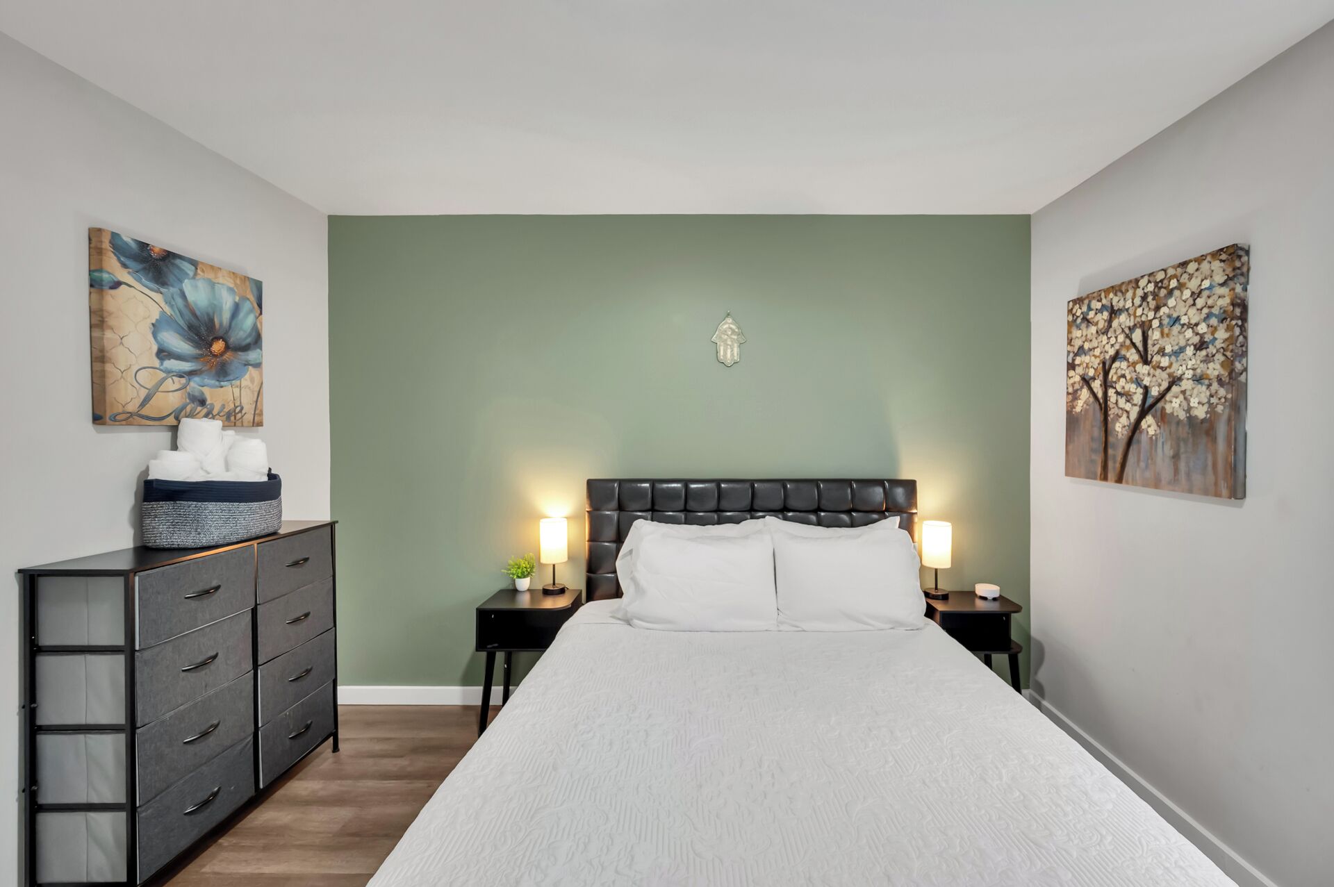 A bedroom with a large bed, two bedside tables with lamps, a dresser, and two wall-mounted paintings.