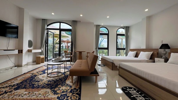 Habitación de hotel con una cama grande, un sofá, una mesita y un televisor. Hay dos ventanas con cortinas, y se ve un balcón a través de ellas.