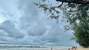 Una playa con gente caminando y parada cerca del agua, un cielo nublado y árboles en primer plano.