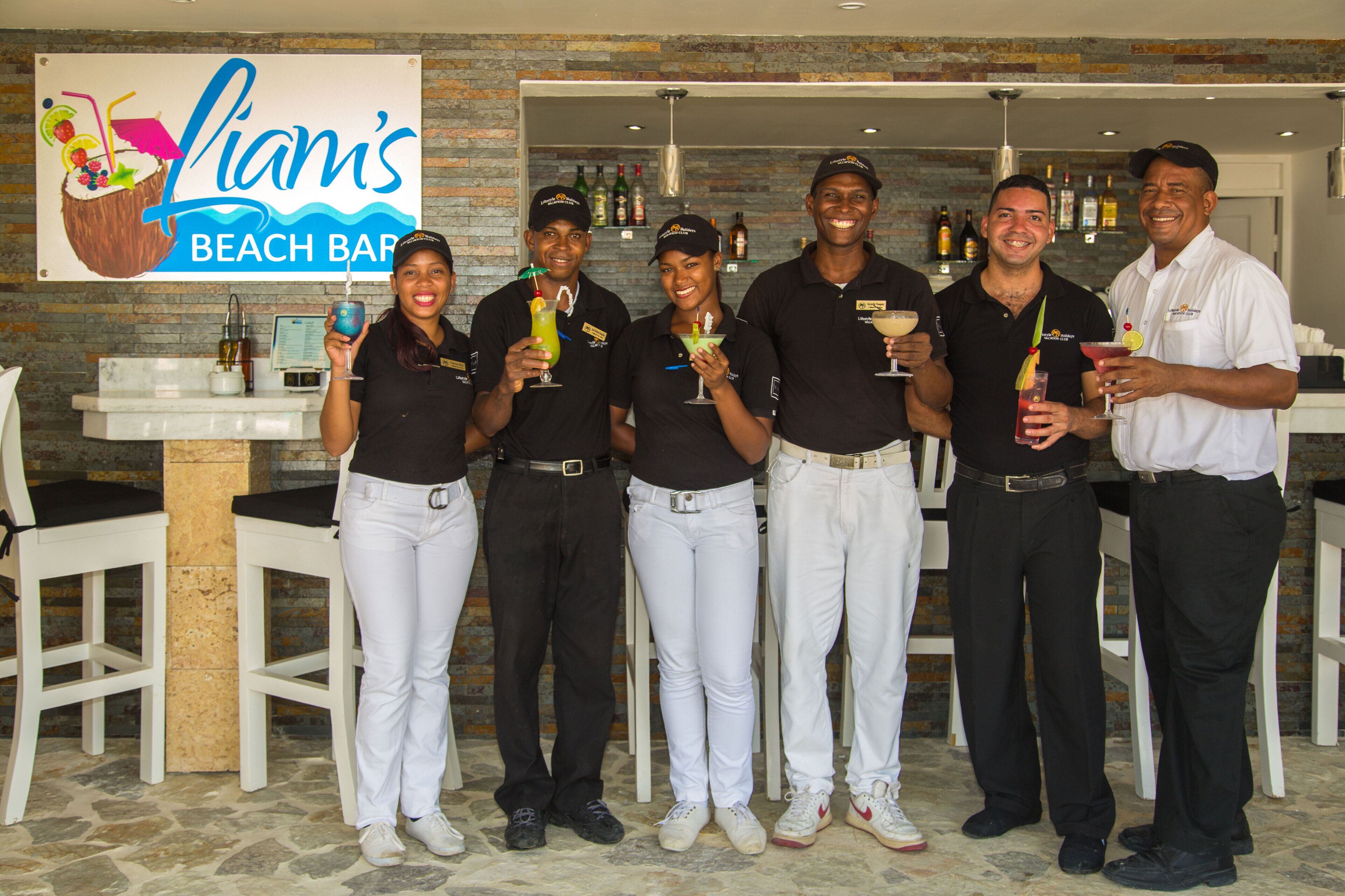 A group of people standing in front of a beach bar sign, holding drinks.