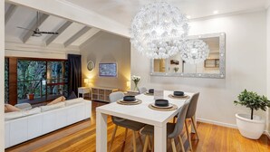 A modern dining area with a white table, grey chairs, and a chandelier. There is a sofa with cushions in the background, a large window with curtains, and a potted plant. - Family-Friendly 3BR Home with Outdoor Living in Ferny Hills (Ferny Hills)