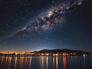 Cielo nocturno con estrellas y la Vía Láctea reflejados en una masa de agua junto a las luces de la ciudad.