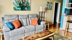 A living room with a grey sofa, colorful cushions, a wooden coffee table, a floor lamp, and a painting on the wall. - Hilo Getaway: 2BR Condo w/ Pool, A/C & Incredible Views (Hilo)