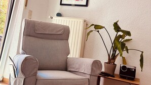 A living room with a grey armchair, a radiator, a framed abstract painting, a potted plant, and a small wooden table with a black device. - Peace & relaxation in the countryside! Mediterranean country house apartment with jacuzzi (Merchweiler)