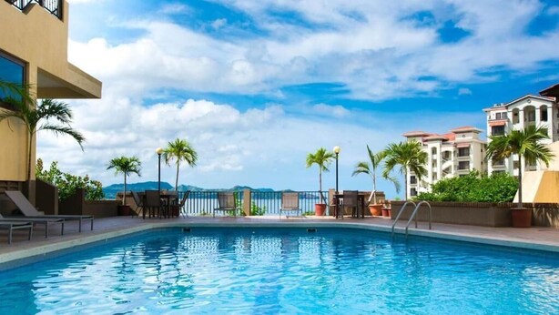 A swimming pool with lounge chairs and palm trees, surrounded by buildings with a view of the mountains. - Studio Apt. Just 5-mins Walk from Flamingo Beach! (Playa Flamingo)