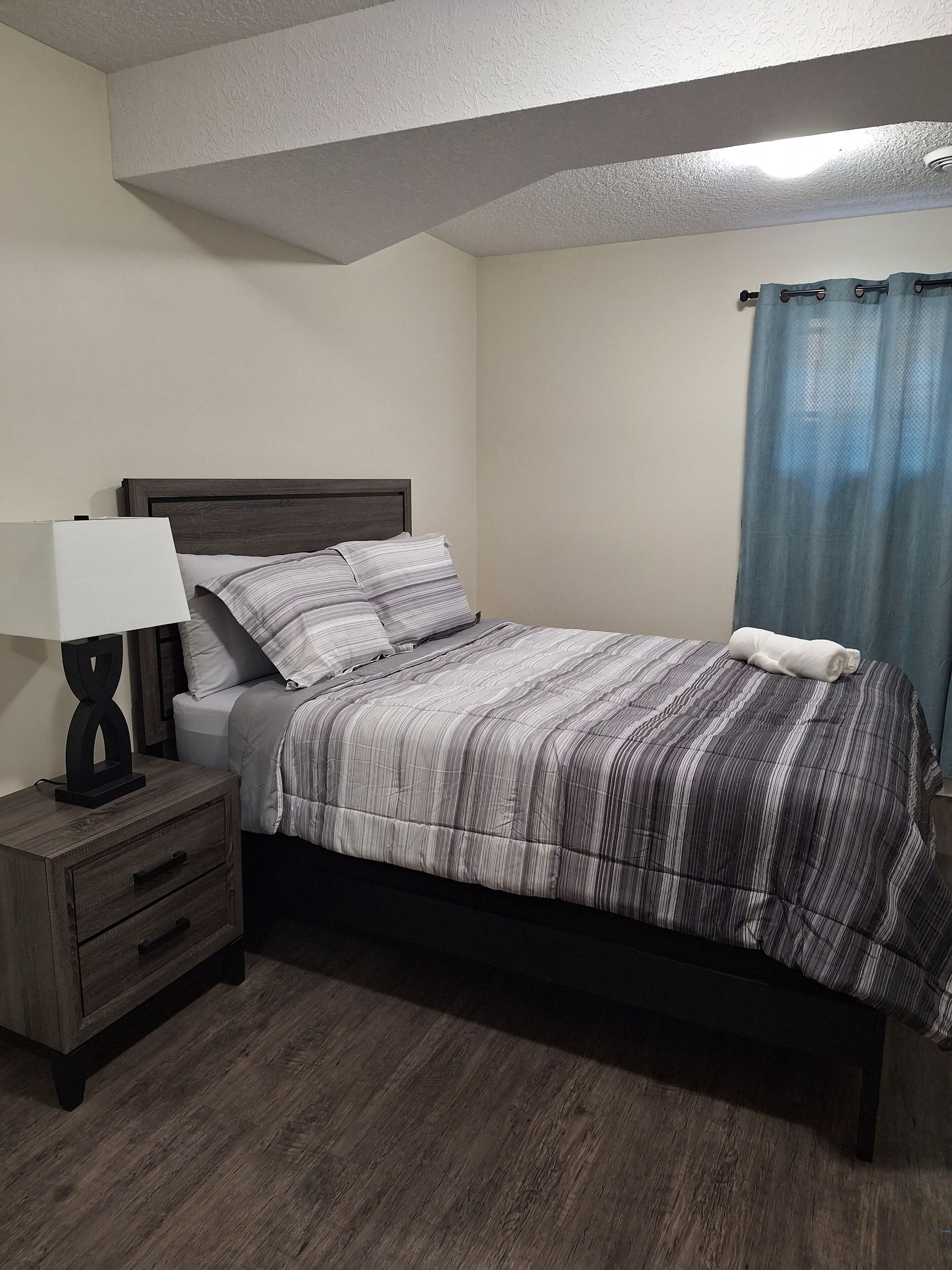Een slaapkamer met een bed, nachtkastje, lamp en gordijn.