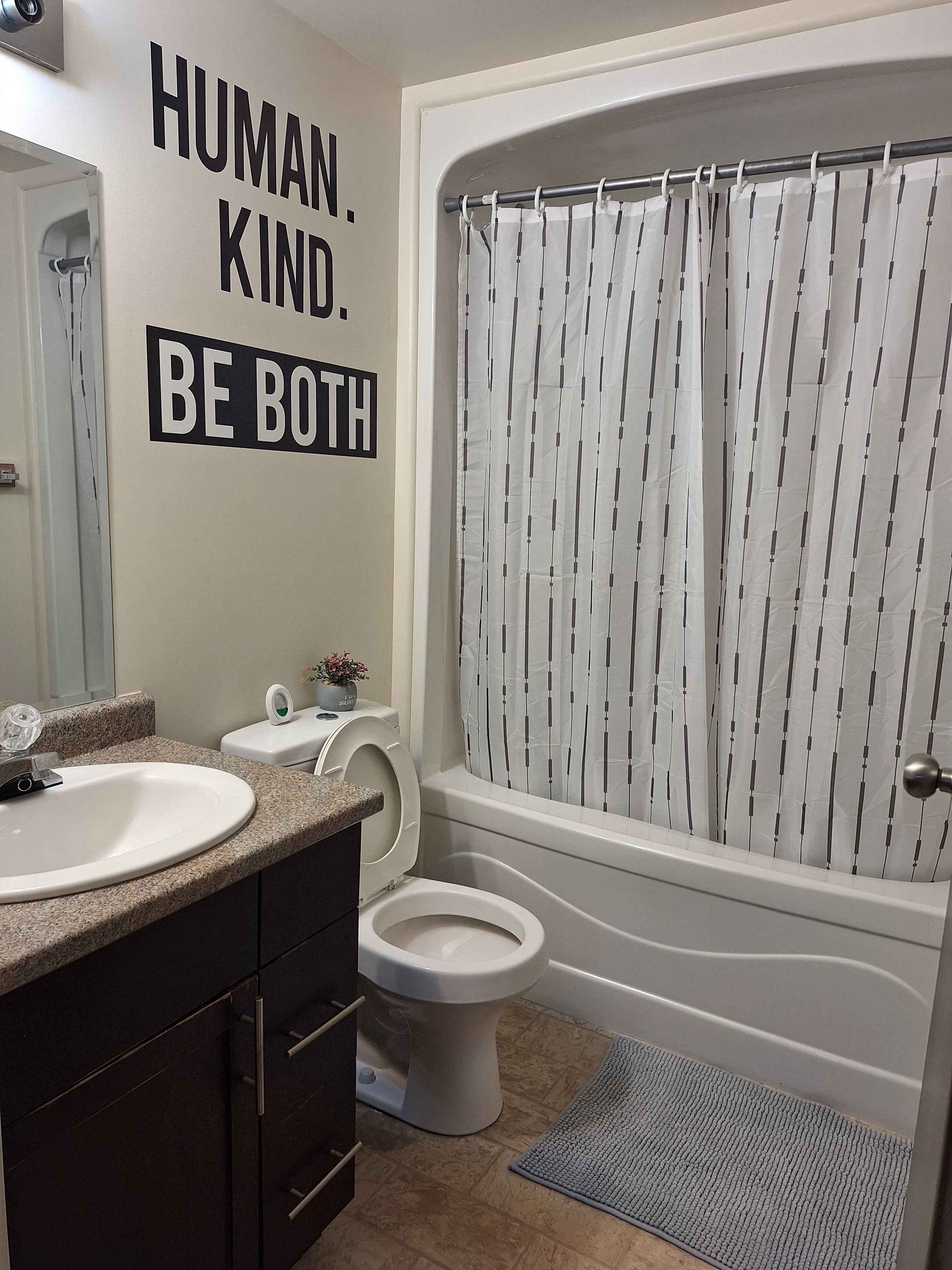 Ein Badezimmer mit Waschbecken, Toilette und Badewanne. An der Wand ist ein motivierendes Zitat und auf der Ablage steht eine kleine Pflanze.