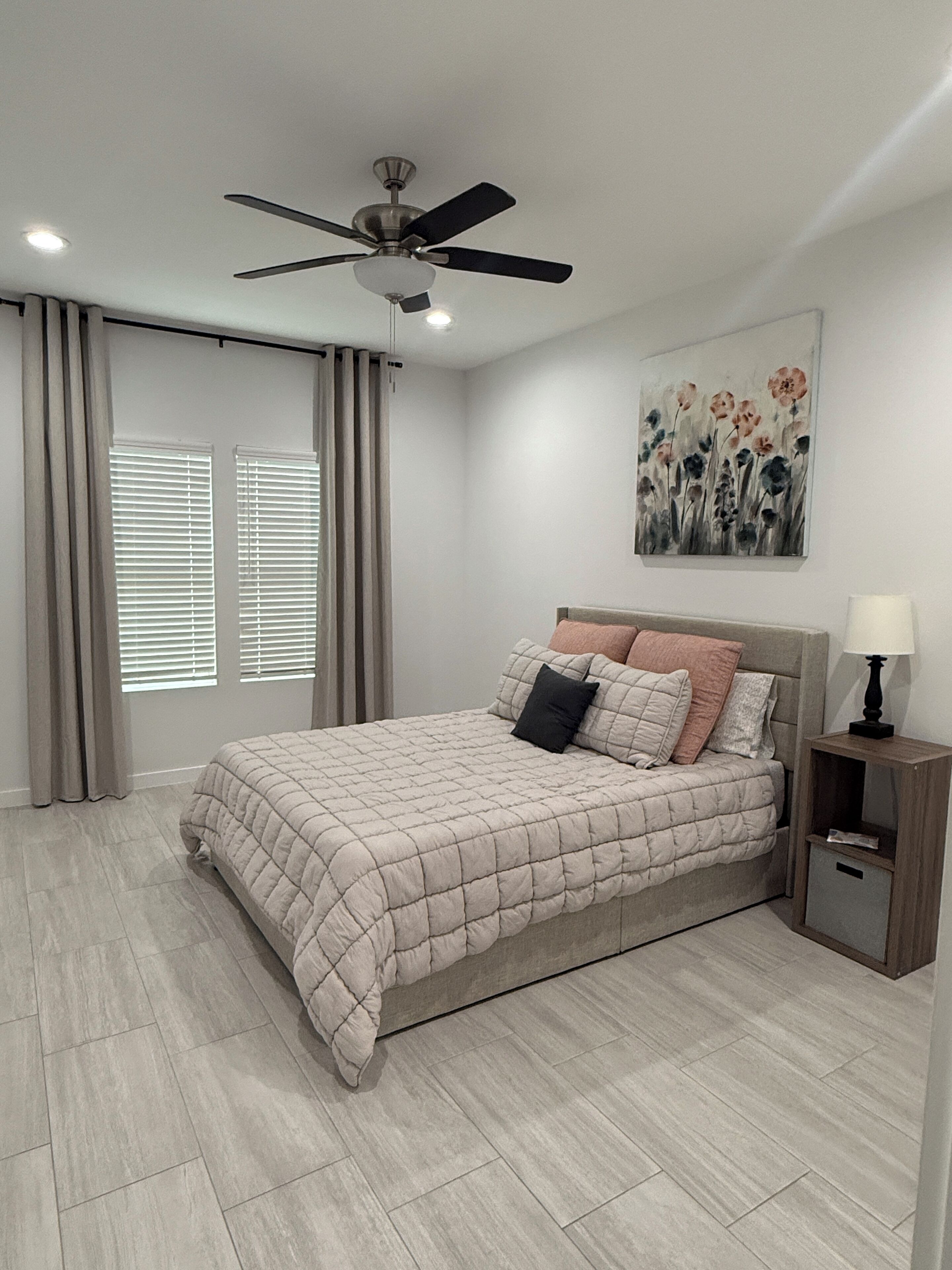 Un dormitorio con una cama, un ventilador de techo, una mesita de noche, una lámpara y un cuadro en la pared.
