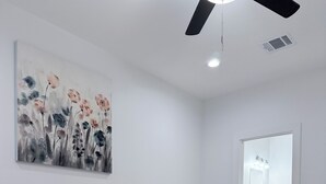 Et moderne soveværelse med en stor seng, et natbord, en lampe, ventilator i loftet, et billede på væggen, og et badeværelse kan ses gennem en åben dør.