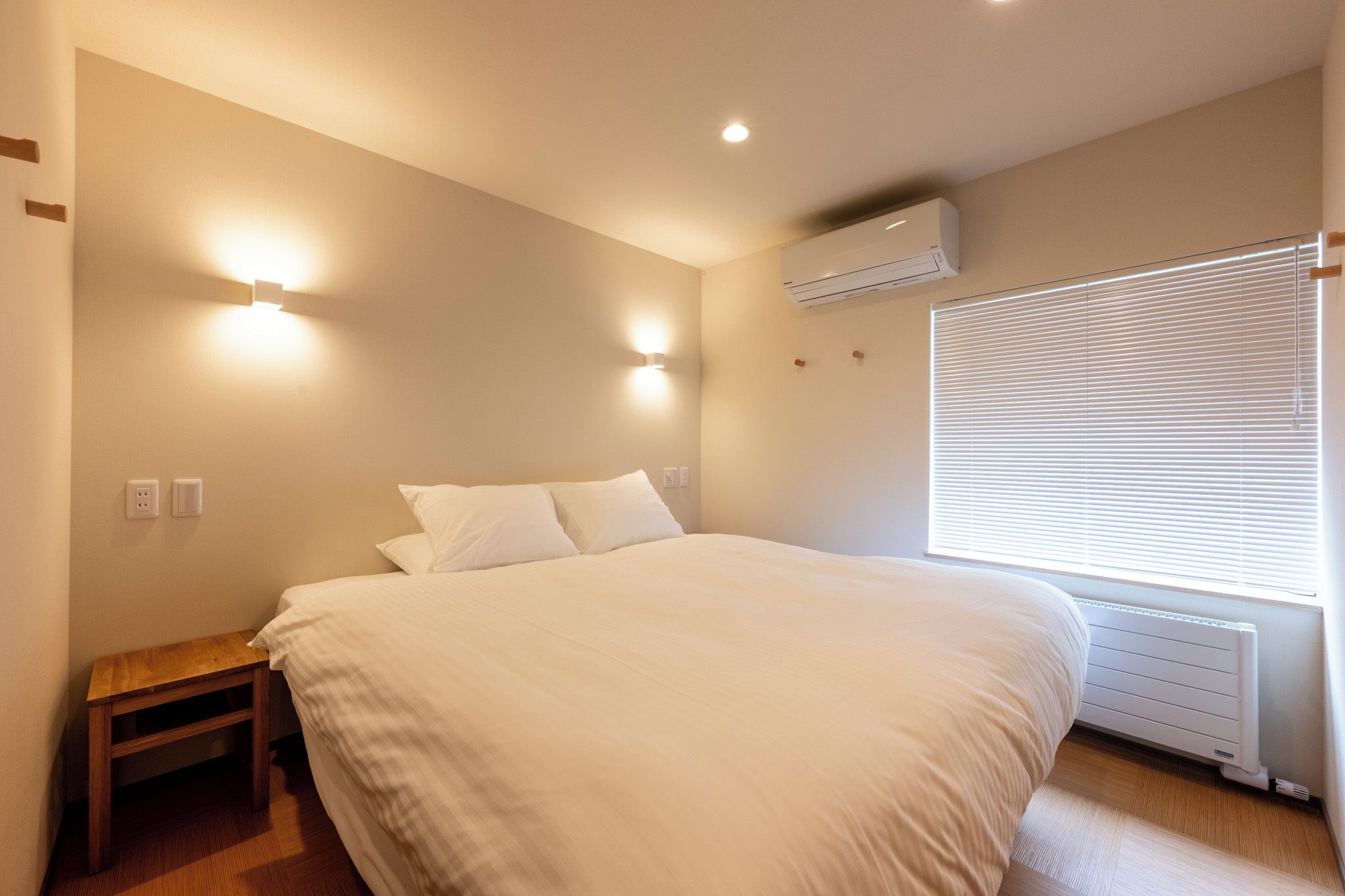 A bedroom with a bed, bedside table, wall-mounted light, air conditioning unit, and a window with blinds.