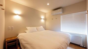 A bedroom with a bed, bedside table, wall-mounted light, air conditioning unit, and a window with blinds.