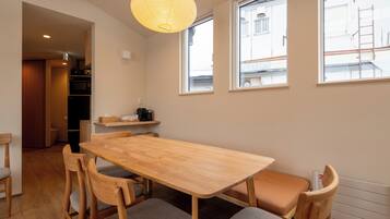 A dining area with a wooden table and chairs, a hanging light fixture, and a view of the street through windows.