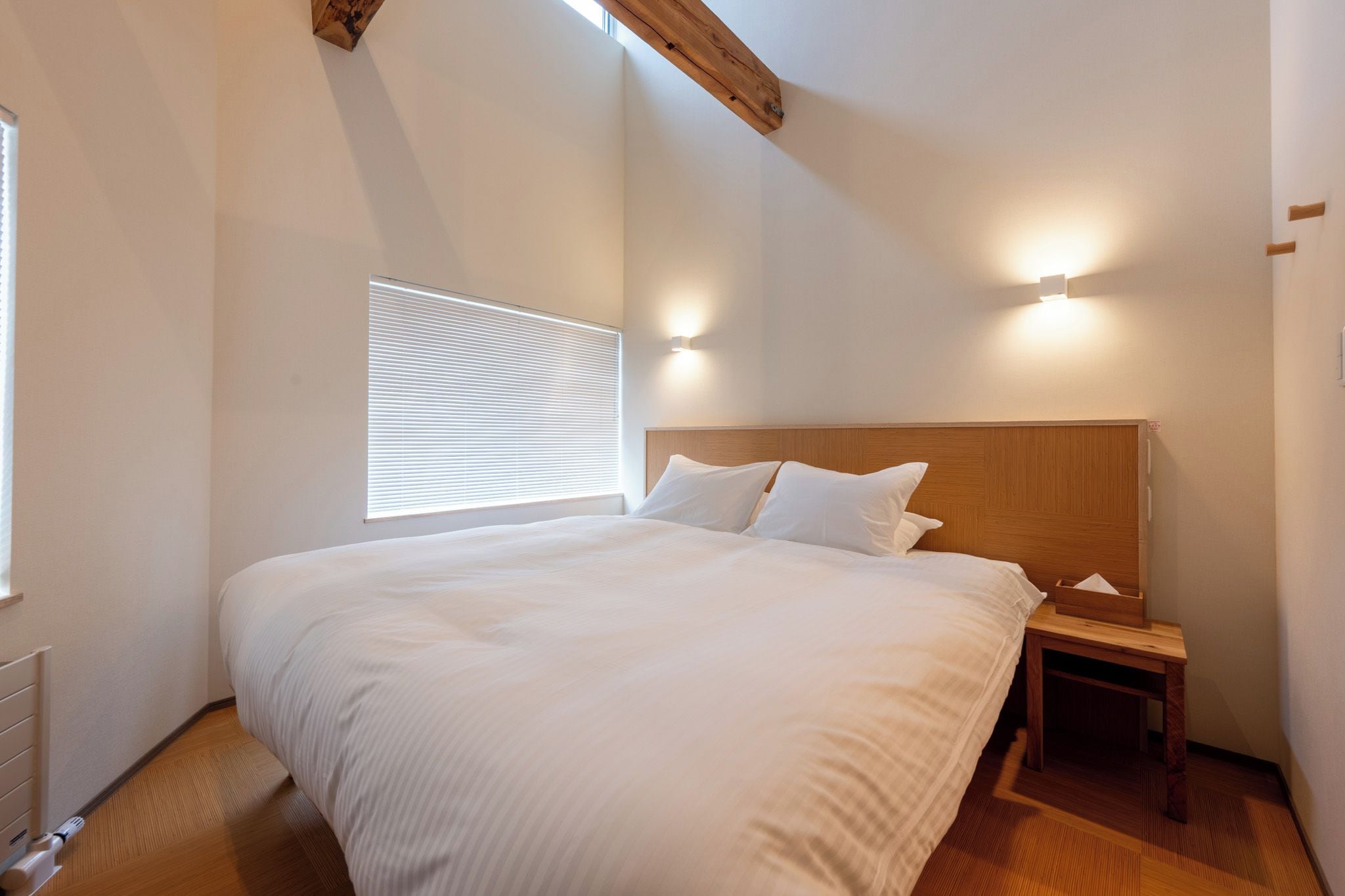 A bedroom with a large bed, a nightstand, a wooden headboard, a window with blinds, and a wall-mounted light fixture.