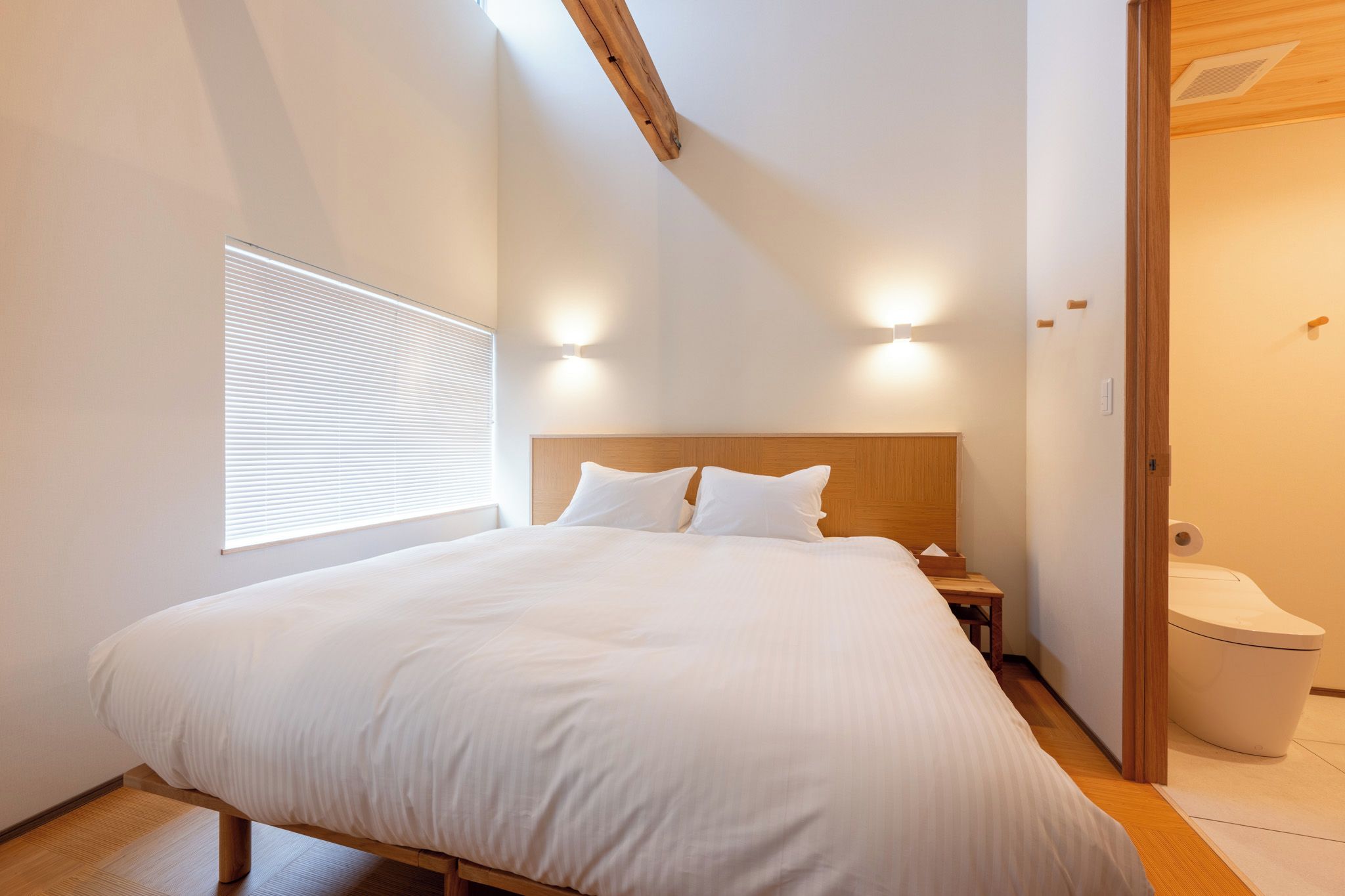 Une chambre à coucher comprenant un lit, des tables de chevet, une tête de lit en bois, une fenêtre avec des stores et une porte donnant sur une salle de bain.