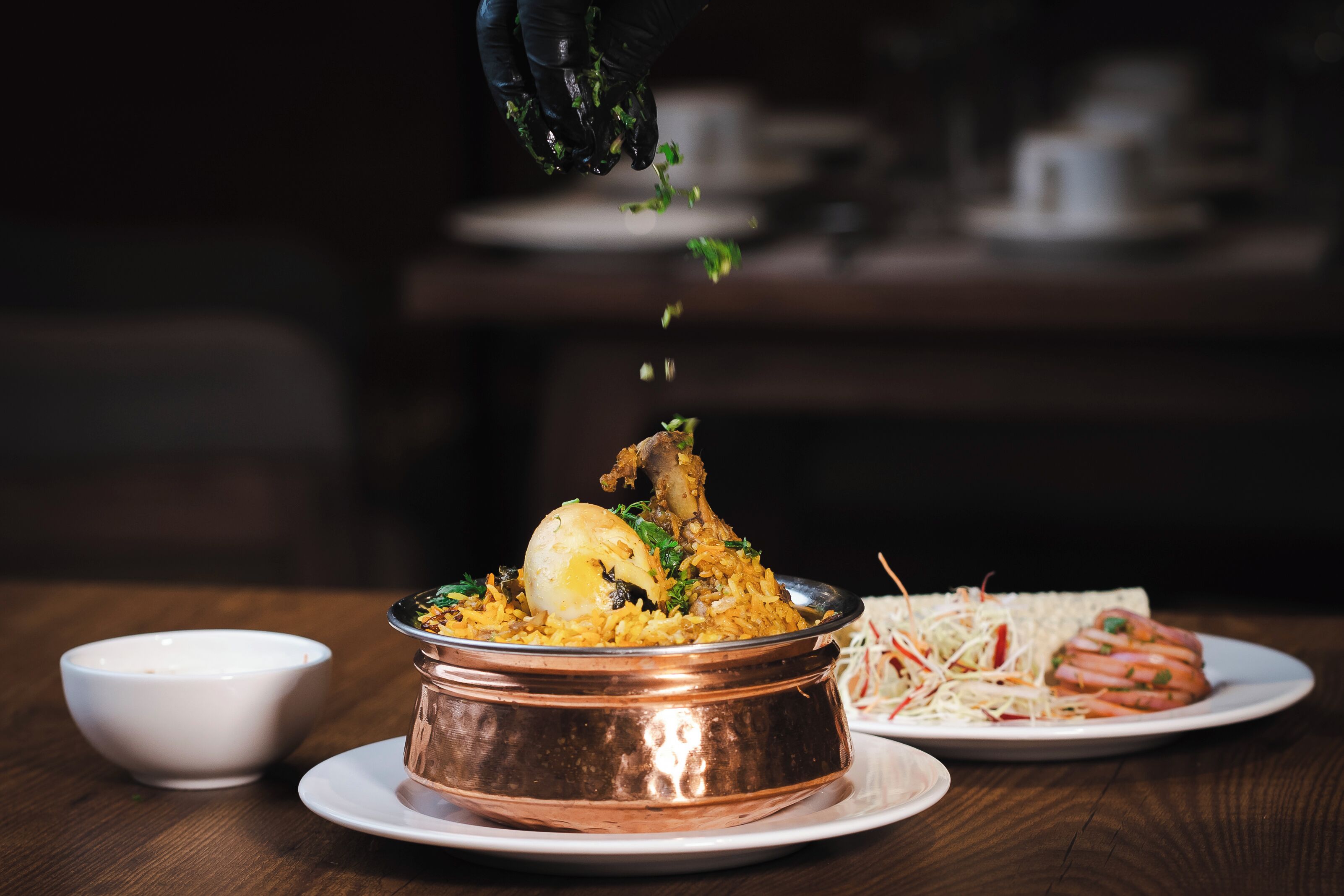 Un tazón de cobre con biryani, una pata de pollo entera, adornado con hierbas, y una ensalada como acompañamiento sobre una mesa de madera.