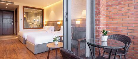 A hotel room with a large bed, a small table with a plant, wicker chairs, and a glass partition.