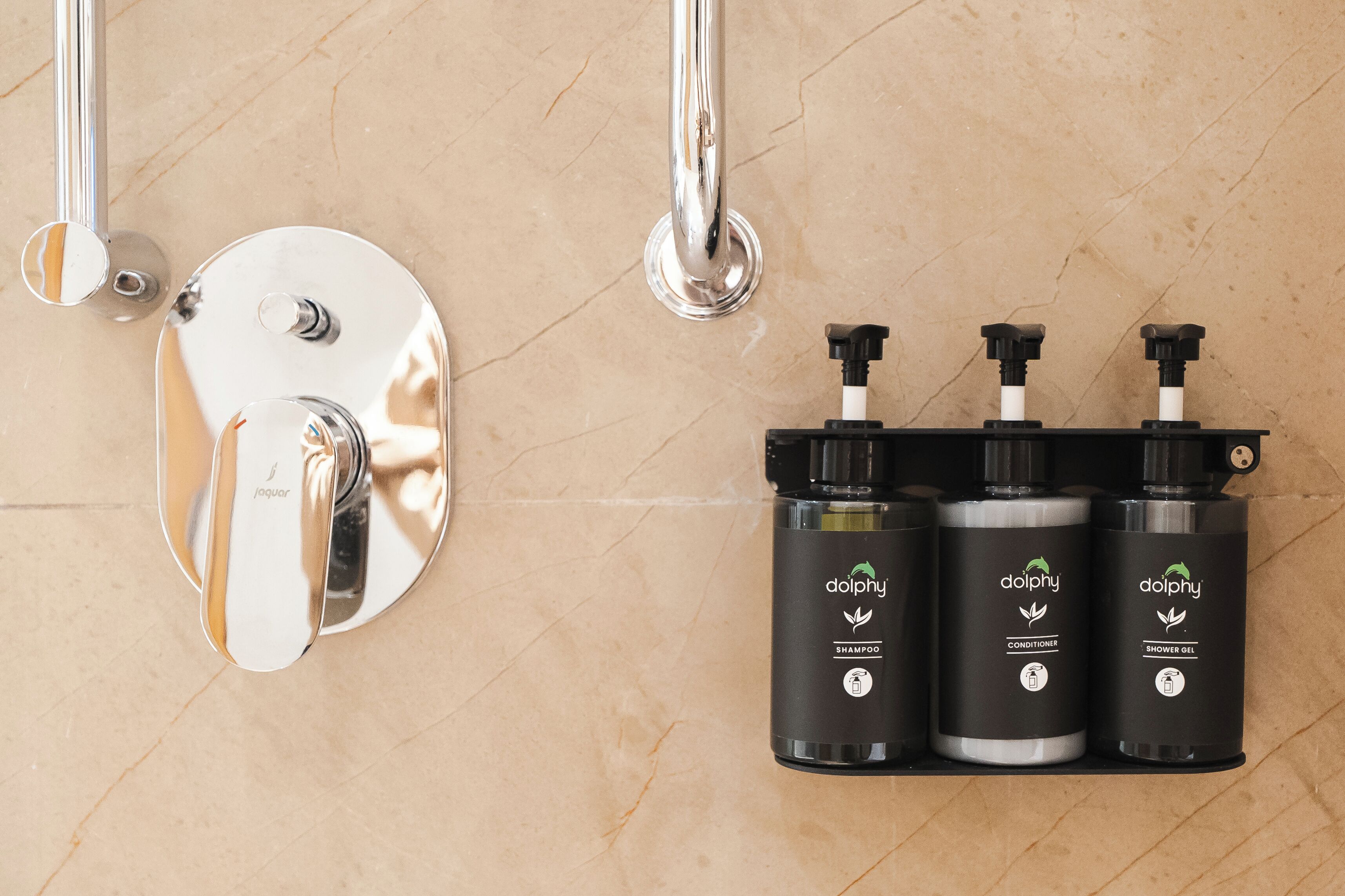 A bathroom with a chrome showerhead, a wall-mounted soap dispenser, and three bottles of Dolphin brand toiletries.
