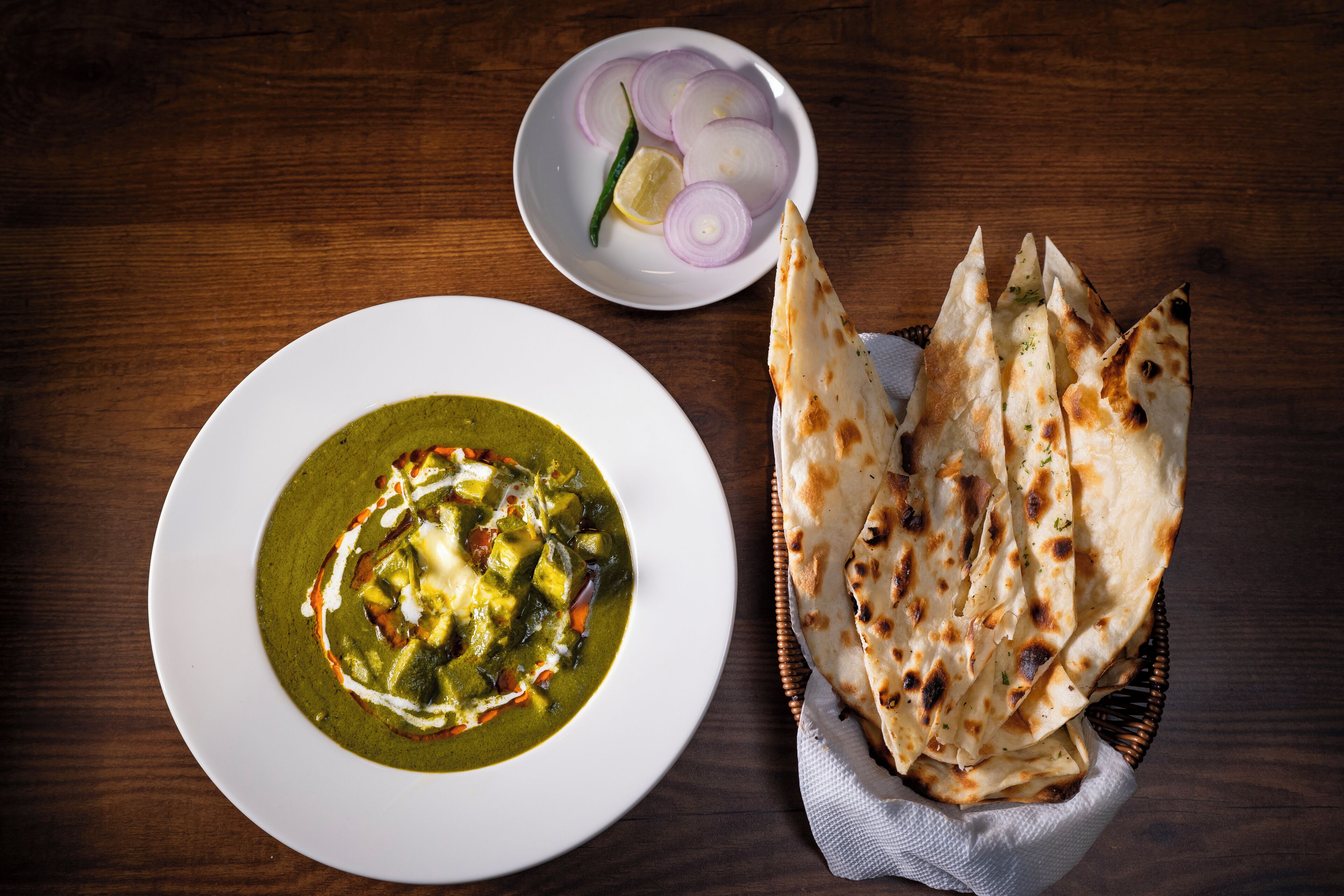 A plate of green curry with naan bread and a bowl of sliced onions and a lemon.