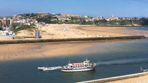 Un pueblo costero con una playa de arena, un barco llamado 'actilur' en el agua y edificios al fondo.