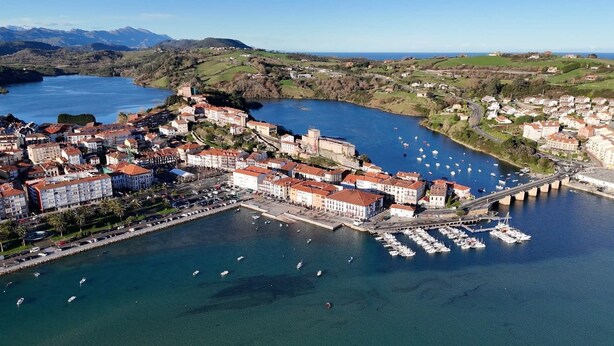 Vista aérea de un pueblo costero con un castillo histórico, un puerto deportivo y edificios residenciales.