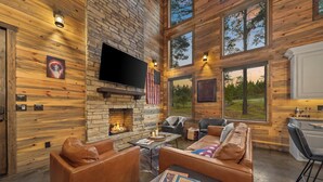 A cozy cabin interior with a stone fireplace, leather seating, and large windows offering views of the outdoors. - Cab'n America - Pets/ Giant Jenga/ Hot tub/ Fire Pit/ Horseshoes (Broken Bow)
