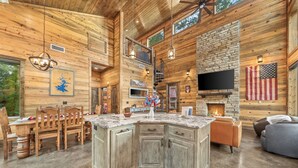 A spacious living area with a large wooden dining table, a stone fireplace, and a television mounted on the wall. - Cab'n America - Pets/ Giant Jenga/ Hot tub/ Fire Pit/ Horseshoes (Broken Bow)