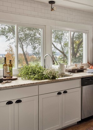 A kitchen with white cabinets, a stainless steel dishwasher, a window with a view of trees, and a countertop with kitchen items.