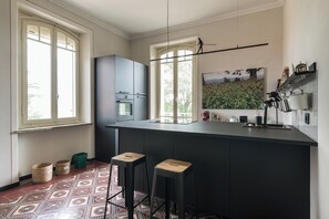 A modern kitchen with a central island, two bar stools, a painting, and a view through the windows.