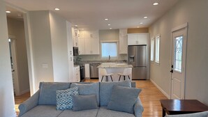 A modern living room with a grey sofa, a coffee table, and a small wooden side table. A kitchen with white cabinets, a refrigerator, and a dining area with two white chairs is visible in the background. - Immaculate and private 2-bedroom house, close to everything, pet friendly (Central Point)