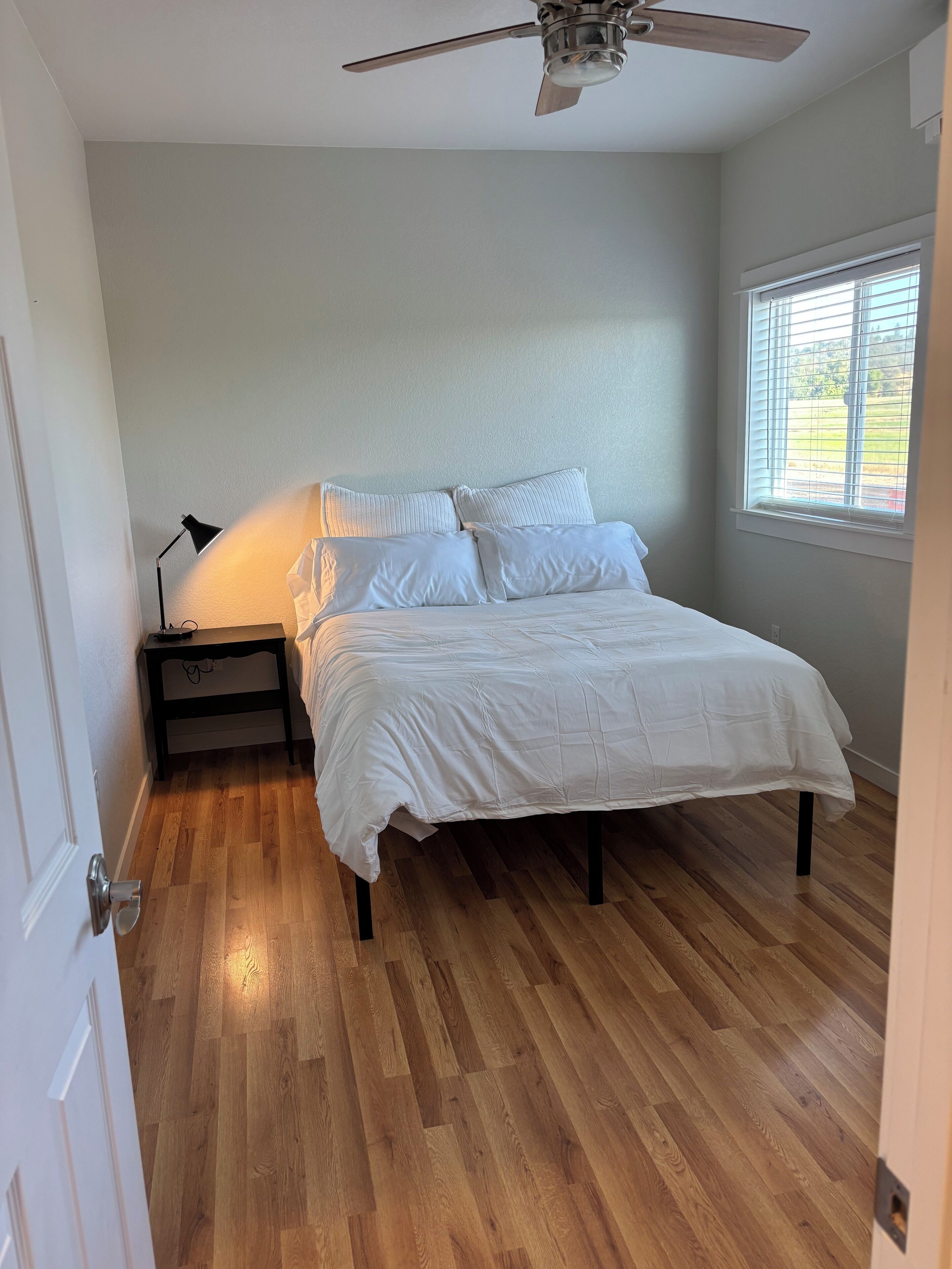 Une chambre à coucher avec un lit, une table de chevet, un ventilateur de plafond et une fenêtre avec des stores.