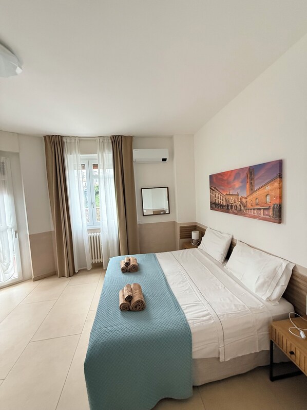 A hotel room with a bed, a bedside table, a wall-mounted air conditioner, a window with curtains, and a painting on the wall.