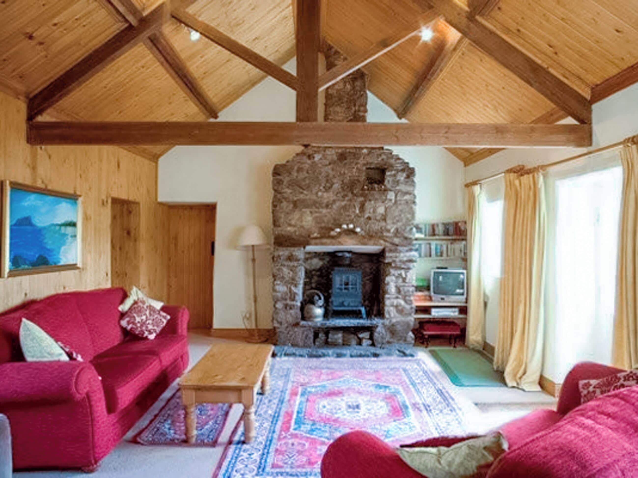A cozy living room with a stone fireplace, wooden beams, a red sofa, and a television.