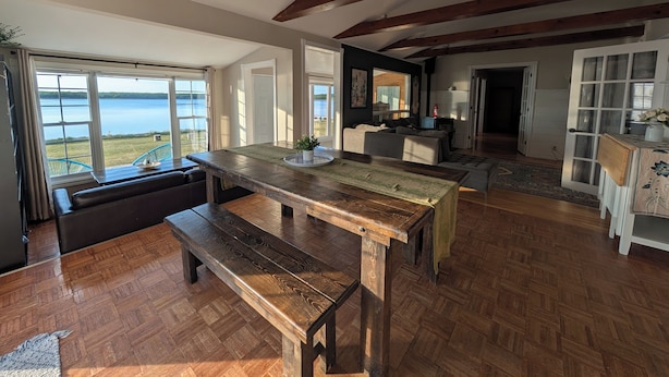 A spacious living area with a large wooden table, a bench, and a view of a lake through open doors.