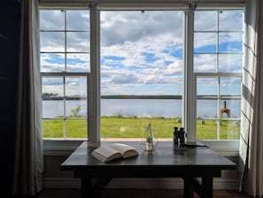 A room with a large window overlooking a lake, a wooden table with a book and a plant, and a tripod with cameras.