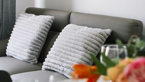 Ein modernes Wohnzimmer mit einem grauen Sofa, zwei weißen Kissen mit Struktur, einem gerahmten abstrakten Gemälde und einem Glastisch mit Blumen in einer Vase.