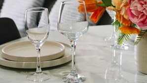 Ein Esstisch mit zwei Weingläsern und Tellern, eine Vase mit Blumen und eine Couch im Hintergrund.